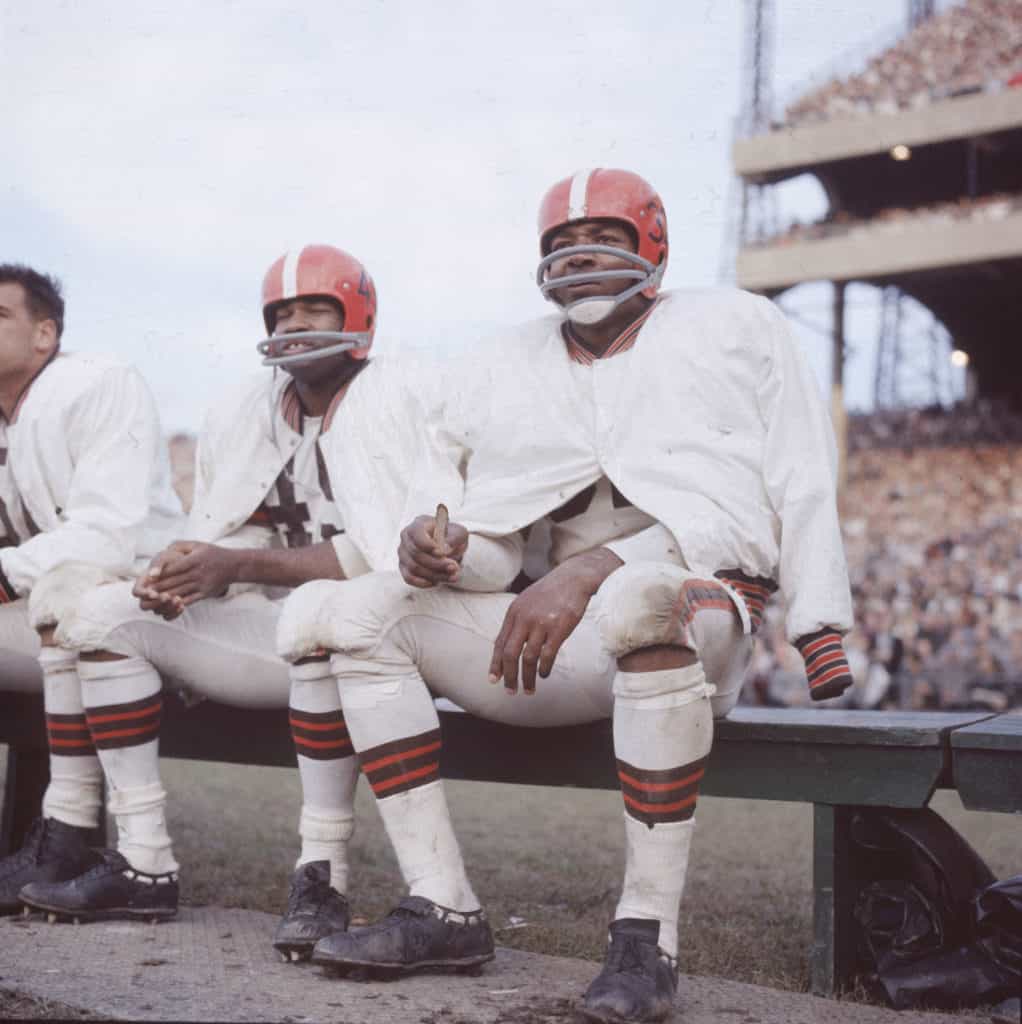 American football player Jim Brown sits on the bench during a game, wearing his helmet and uniform for the Cleveland Browns, circa 1960.