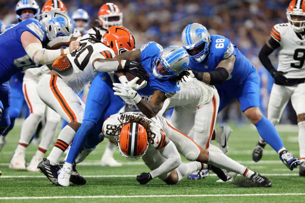 DETROIT, MICHIGAN - SEPTEMBER 28: Jahmyr Gibbs #0 of the Detroit Lions is tackled by Devin Bush #30 and Ronnie Hickman Jr. #33 of the Cleveland Browns during the fourth quarter at Ford Field on September 28, 2025 in Detroit, Michigan.