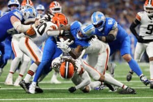 DETROIT, MICHIGAN - SEPTEMBER 28: Jahmyr Gibbs #0 of the Detroit Lions is tackled by Devin Bush #30 and Ronnie Hickman Jr. #33 of the Cleveland Browns during the fourth quarter at Ford Field on September 28, 2025 in Detroit, Michigan.